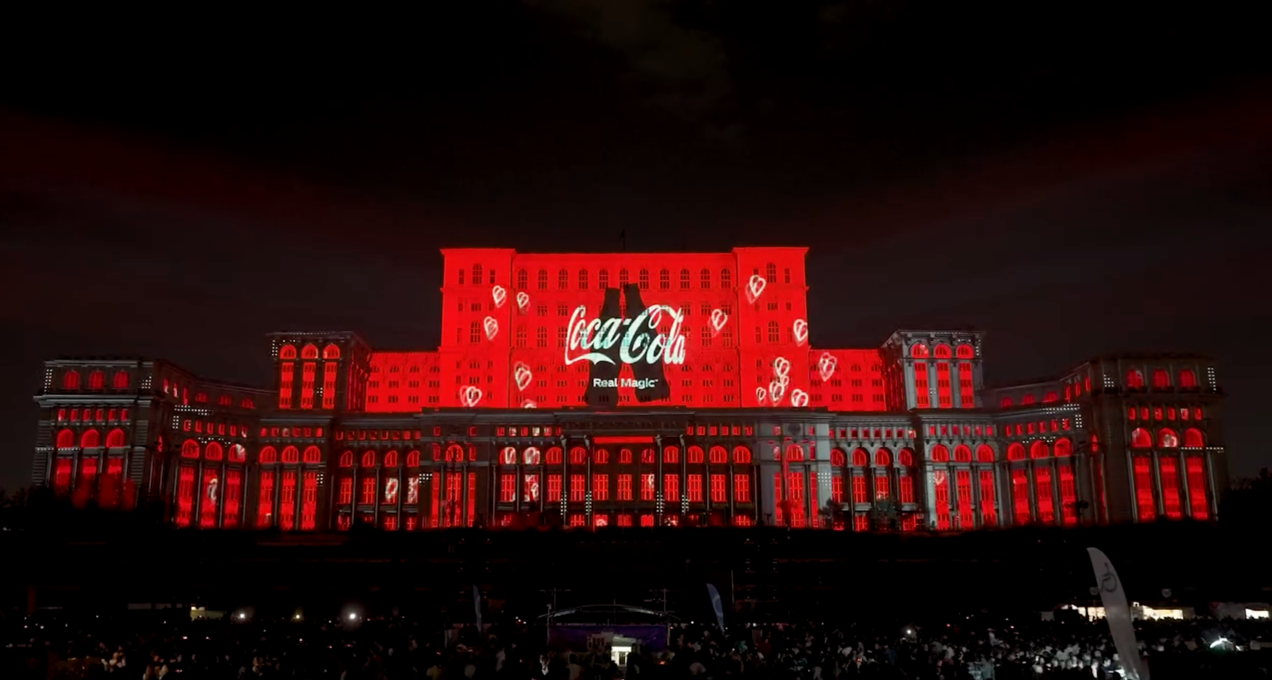 A large building is illuminated with red lights, displaying a projection of the Coca Cola logo along with hearts and bottles on its facade at night.