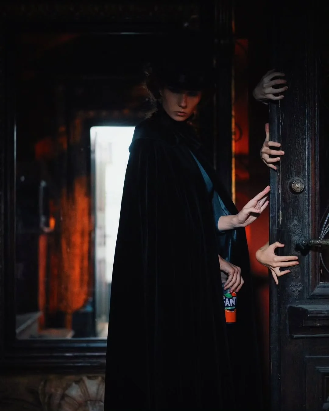 A woman dressed as a witch wearing a black cape and hat, standing in a dimly lit room, holding onto an open door with multiple hands reaching out from behind it.