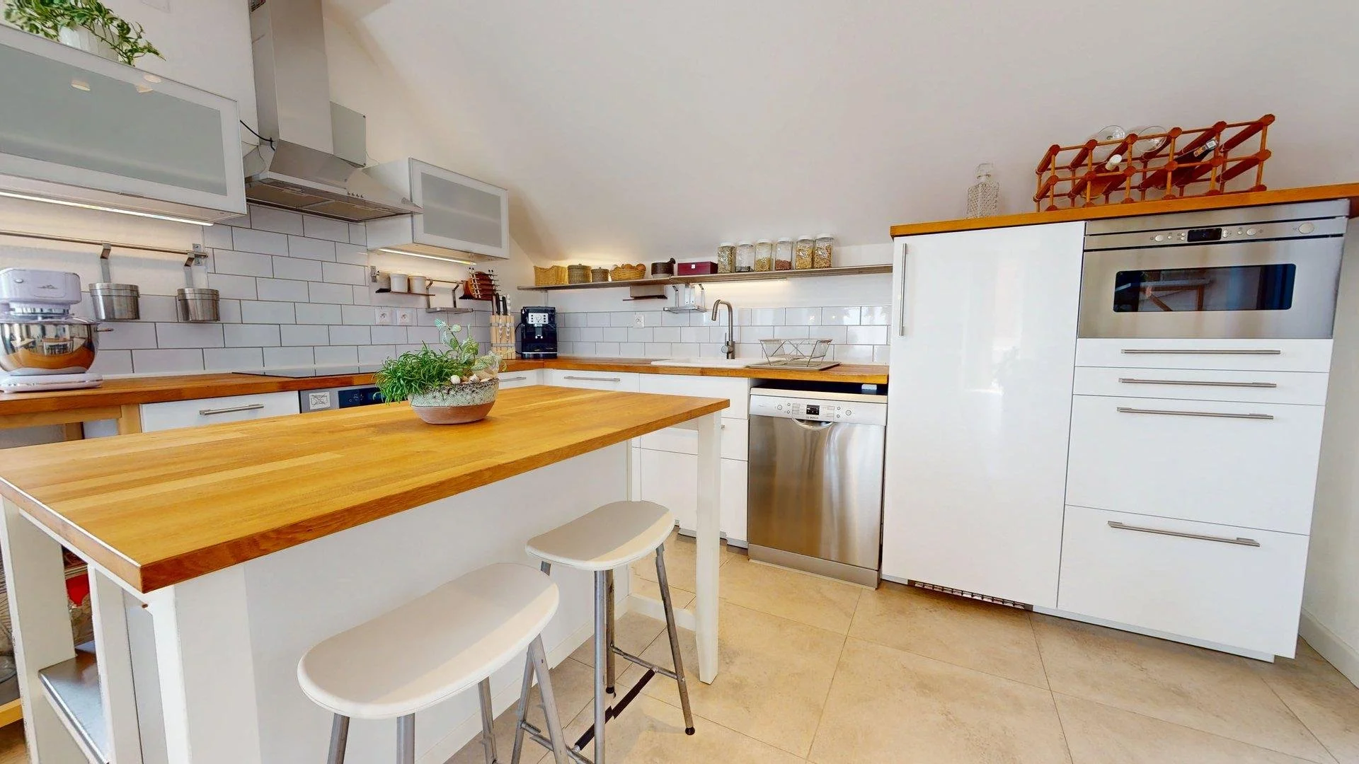 Modern kitchen with white cabinets, a wooden countertop, stainless steel appliances including a dishwasher and oven, and a kitchen island with bar stools. White subway tile backsplash and glass jars on shelves.
