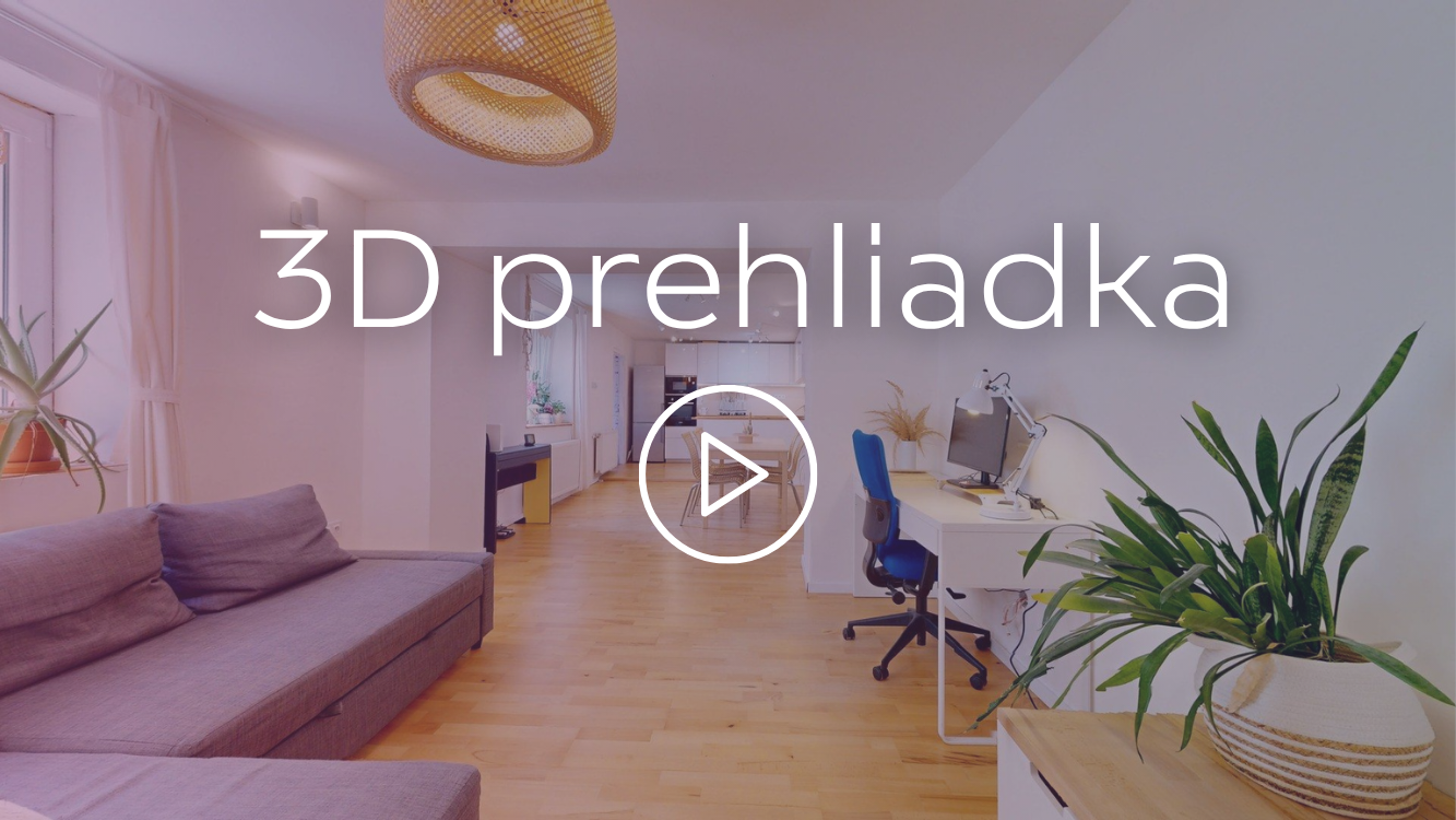 Interior view of a spacious, well-lit apartment with a living room and kitchen area. There is a purple sofa, plants, a computer desk with a chair, and wooden flooring.