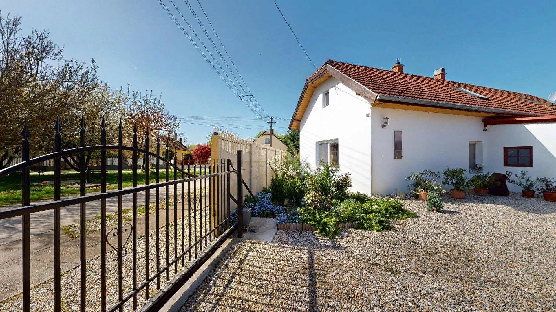 View of a white house with a red tiled roof, surrounded by a gravel yard and potted plants, with a black wrought iron gate at the entrance and a sidewalk with grassy lawn on the other side.