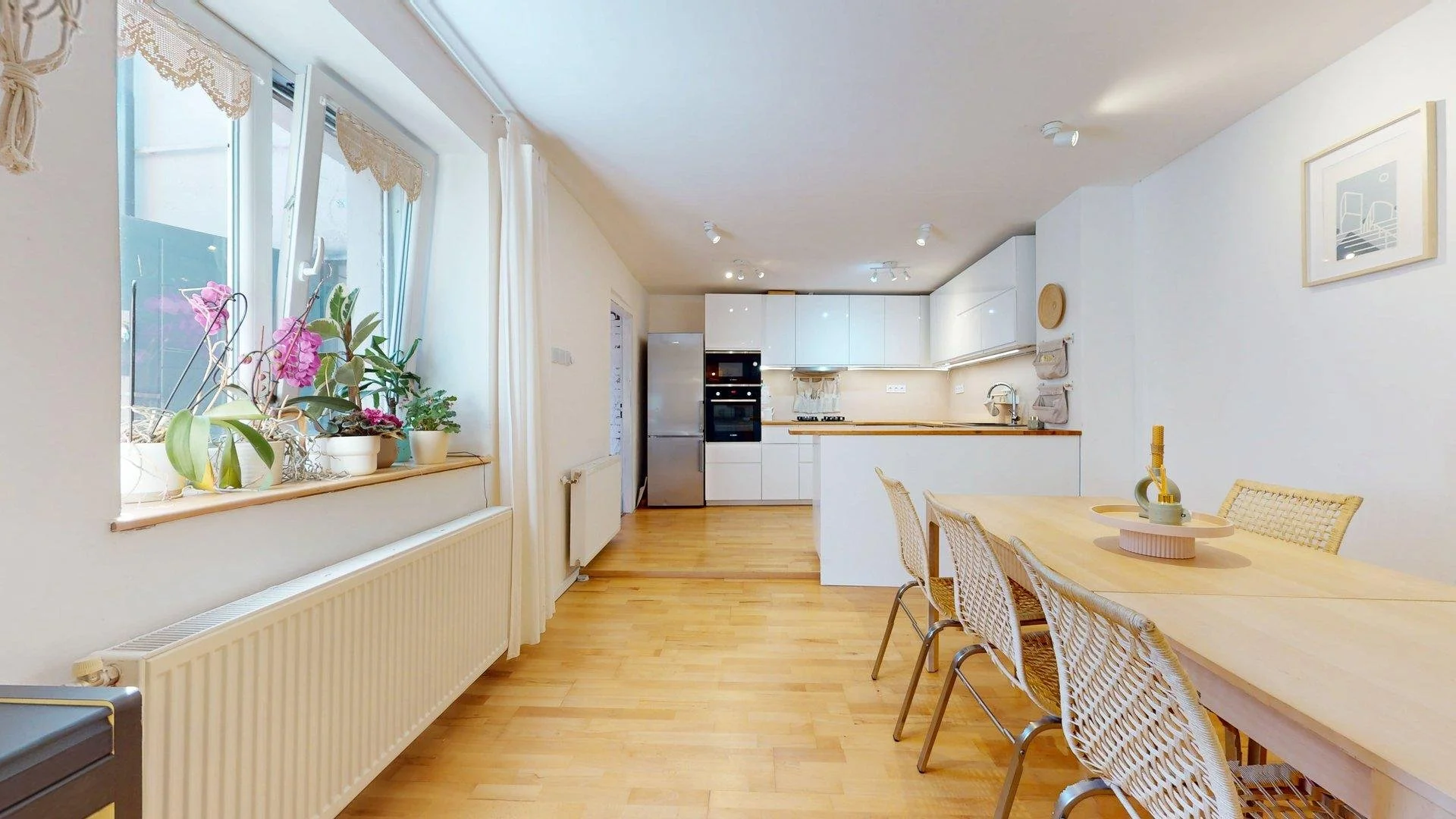 Kitchen with a wooden dining table and six wicker chairs, white cabinets, stainless steel refrigerator, window with plants on the sill, and minimal decor.