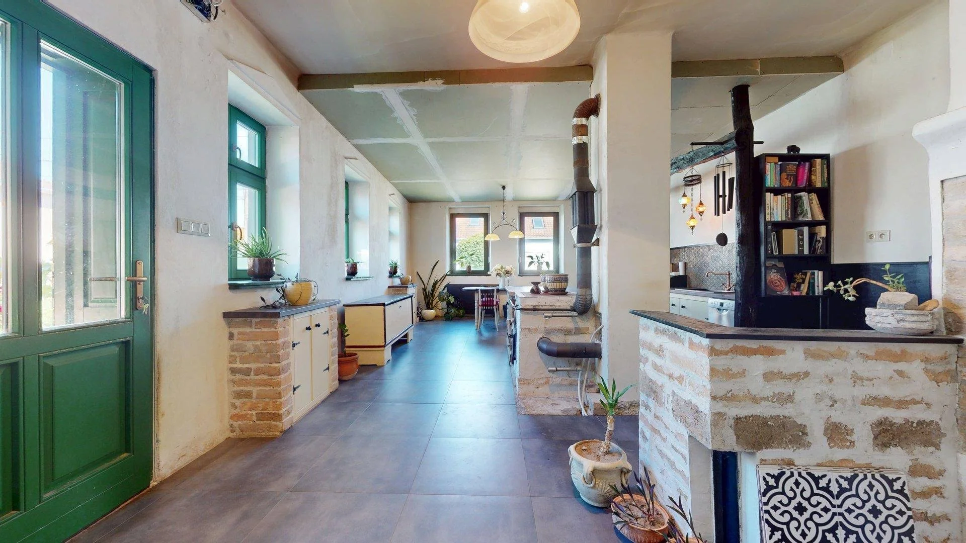 Bright, rustic kitchen with large windows, green door and window frames, brick and plaster walls, and various houseplants. Open space with dining table near the windows, and a black bookshelf with decorative items. Exposed chimney pipe and a mix of modern and vintage decor.