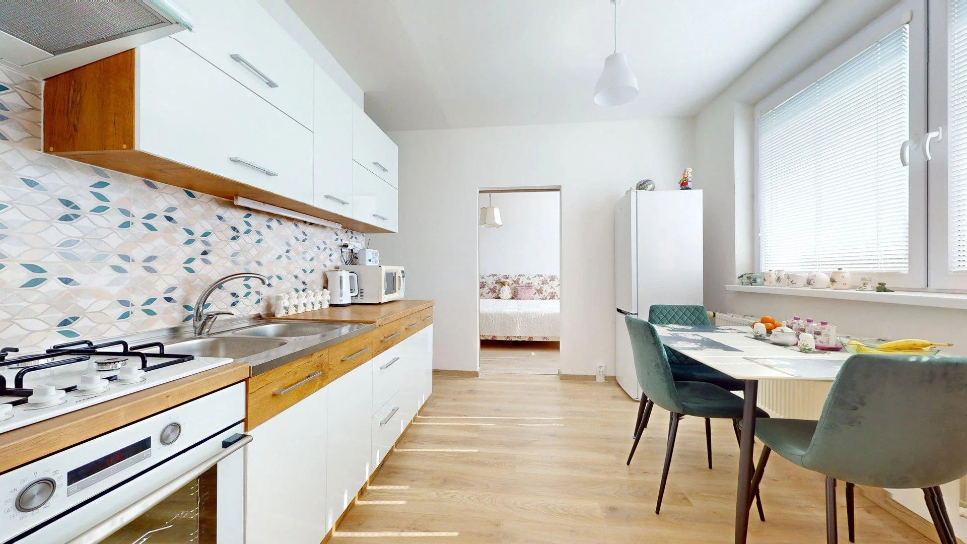 Kitchen with white cabinets, wooden counter, and patterned backsplash. Includes stove, sink, microwave, kettle, and small appliances. Dining area has a table with green chairs and large window with blinds. Doorway leads to a bedroom with bed and floral wallpaper.