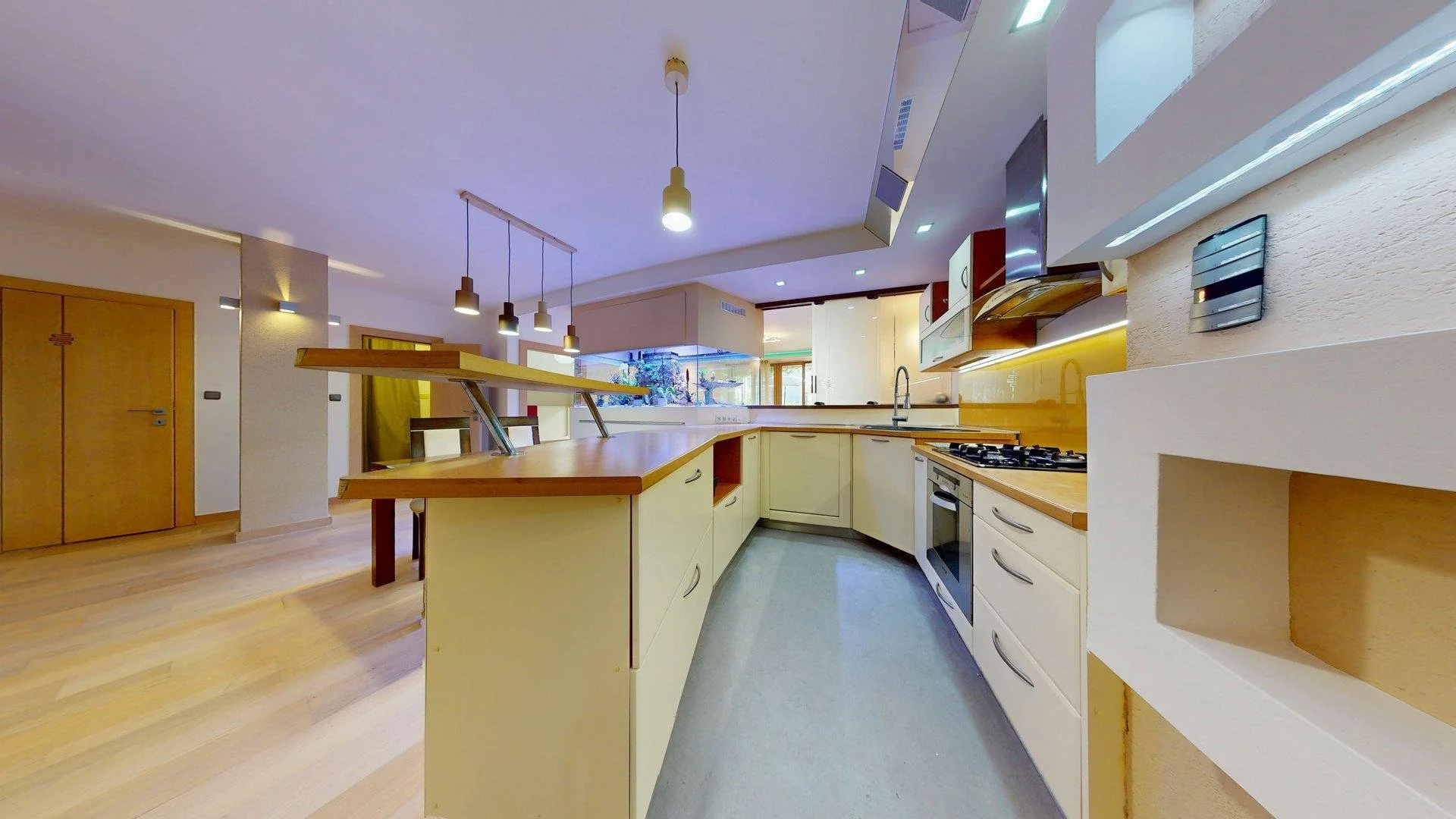 Modern kitchen with cream cabinets and wooden countertops, multiple pendant lights, and a view into the living area with wooden flooring.