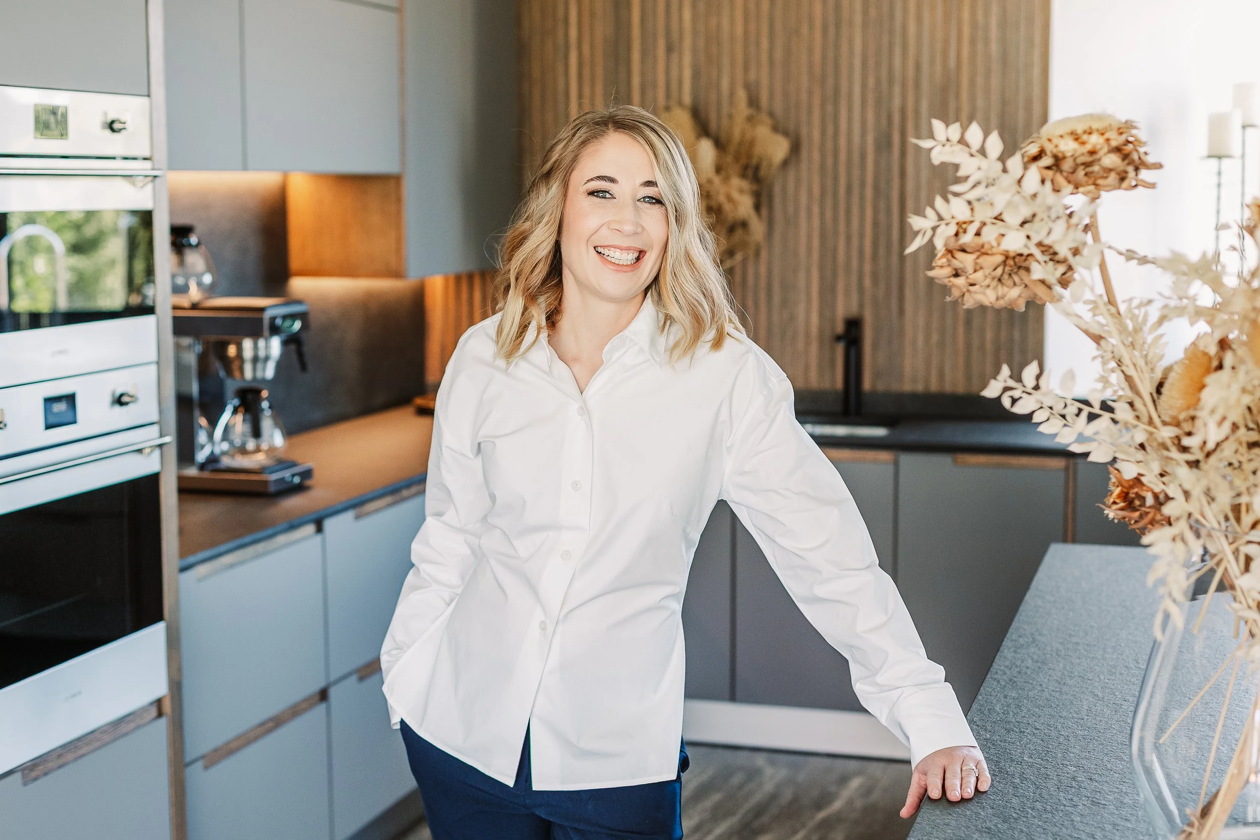 A woman with blonde hair in a white shirt smiling in a modern kitchen.