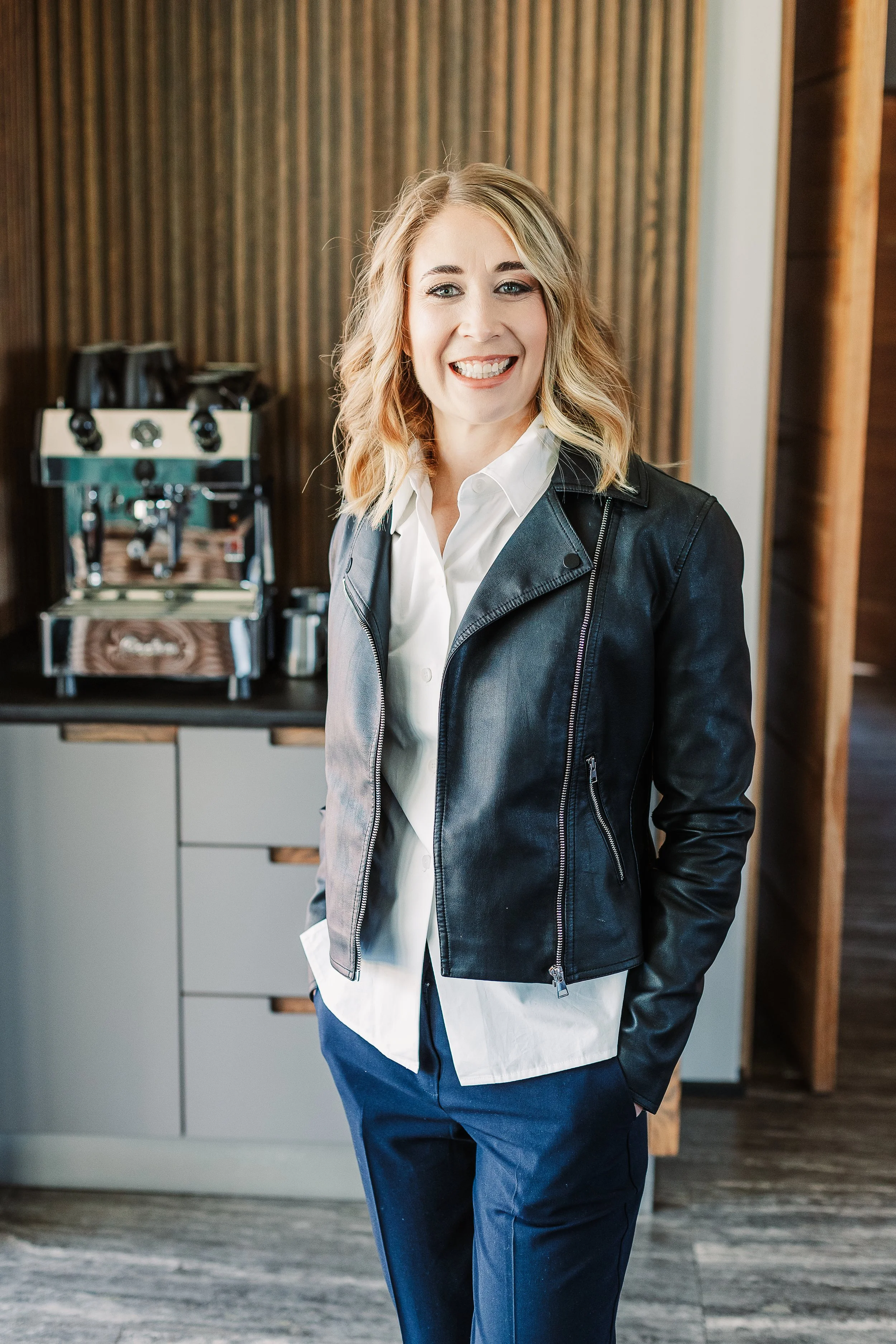 A woman with blonde hair smiling, wearing a black leather jacket, white shirt, and blue pants, standing indoors near a coffee machine.