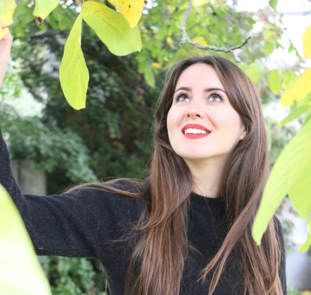 Junge Frau mit braunen Haaren und rotem Lippenstift, die in einem grünen Garten unter Blättern steht.