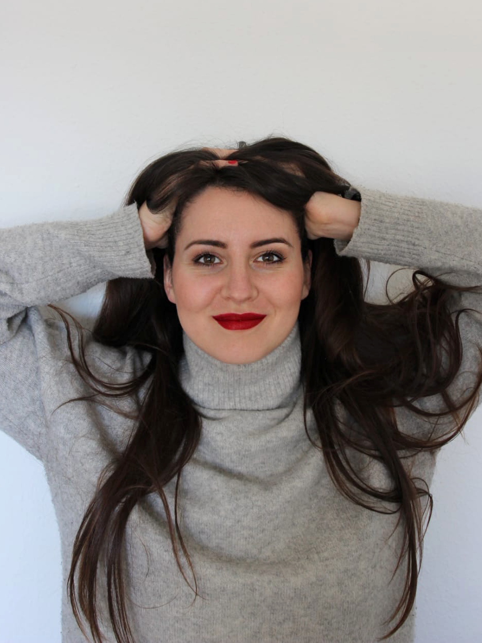 Eine junge Frau mit langem braunem Haar trägt einen grauen Rollkragenpullover und lächelt in die Kamera, während sie ihre Hände in den Haaren hält.