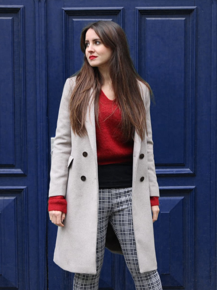 Eine junge Frau mit langen braunen Haaren steht vor einer blauen Tür. Sie trägt einen grauen Mantel, einen roten Pullover und schwarz-weiß karierte Hosen.