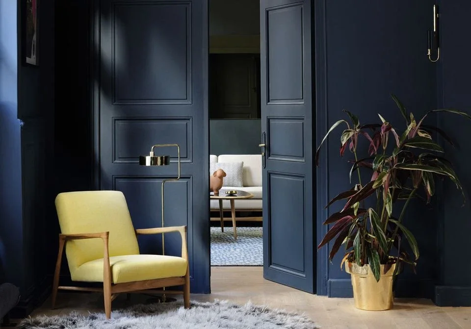 Un coin intérieur avec une porte en bois bleu, un fauteuil jaune, une lampe en métal doré, une plante verte dans un pot doré, un tapis gris et un espace de salon visible à l'arrière.