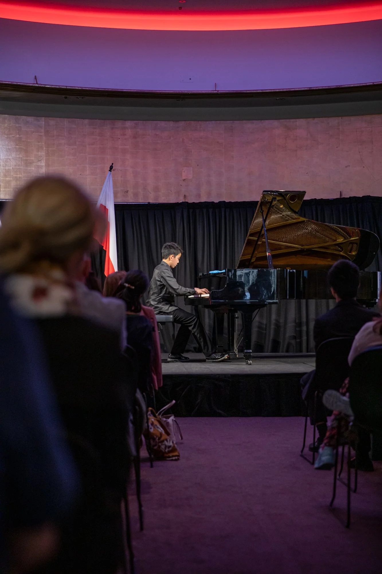 A young man playing a grand piano on a stage with a black curtain backdrop. Audience members are seated facing him, with some visible in the foreground. PolMusicA. Competition. Young talents. Australia. Melbourne, Sydney, Brisbane, Adelaide, Perth.
