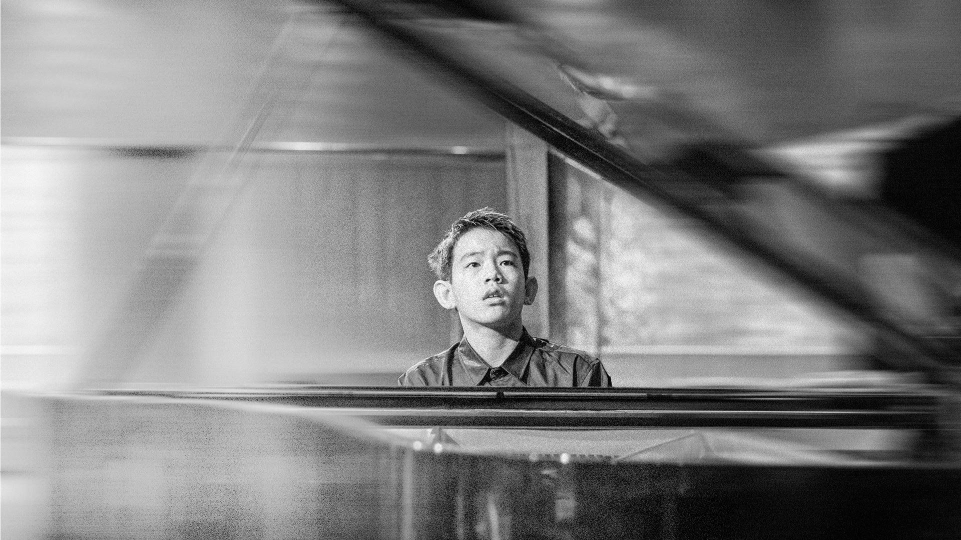 A black and white photo of a young man sitting at a piano with a focused expression on his face. PolMusicA. Competition. Young talents. Australia. Melbourne, Sydney, Brisbane, Adelaide, Perth. Victoria, NSW, Queensland, SA, WA