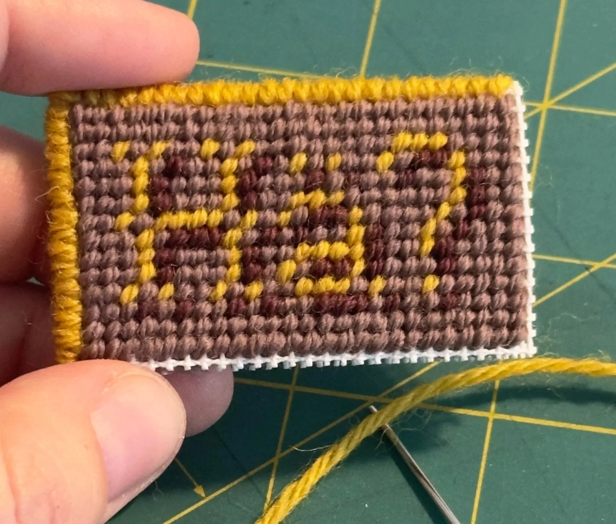 Close-up of a woven patch with a brown and yellow striped pattern, held by fingers, with a green cutting mat with yellow grid lines in the background.