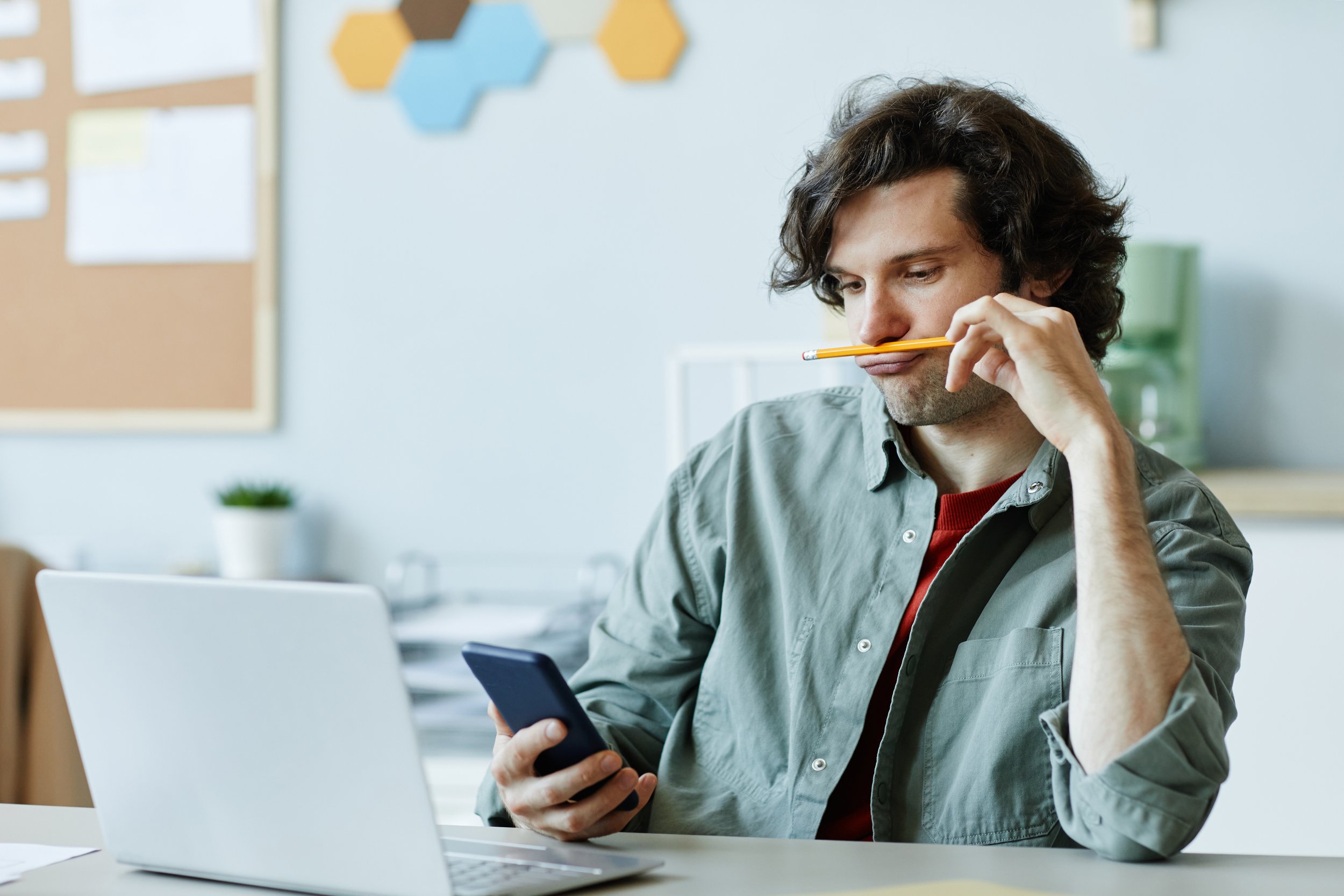 A man with curly dark hair sitting at a desk in a Chicago office, holding a smartphone in one hand and a pencil balanced in his mouth. He appears to be distracted from work and is looking at his phone.
