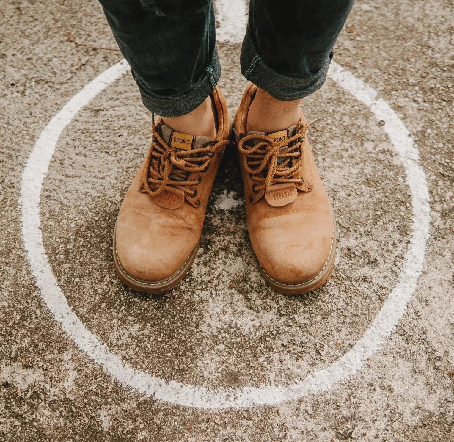 Close-up of tan rugged boots on a person who standing inside a white circle painted on a dirt ground, as if they were stuck.