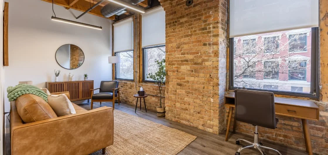 Office space with exposed brick walls, large windows, a leather sofa, a black armchair, a wooden sideboard, a round mirror, and a wooden desk with a rolling chair.