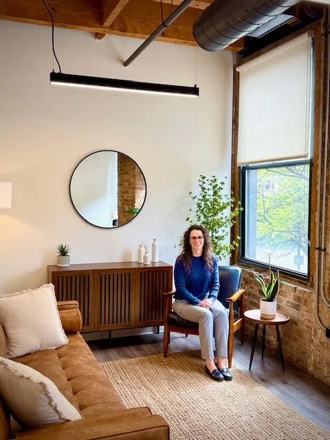 A woman with glasses and curly hair sitting on a wooden chair in a cozy, well-decorated room with large window, potted plants, a mirror, a couch with cushions, and a wooden sideboard.