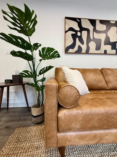 Living room with a large Monstera plant next to a tan leather sofa, a white pillow, wooden side table with electronic devices, a woven basket planter, a textured rug, and a black and white abstract wall art.