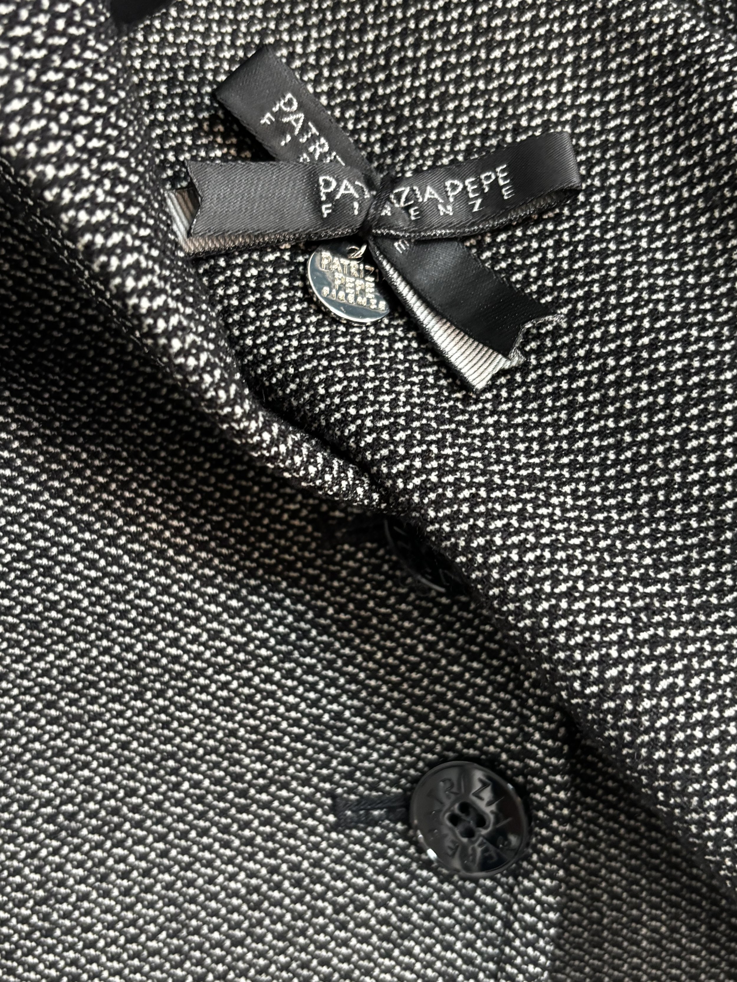 Close-up of Patrizia Pepe Firenze designer blazer featuring a branded black ribbon bow, metallic logo charm and textured grey woven fabric.