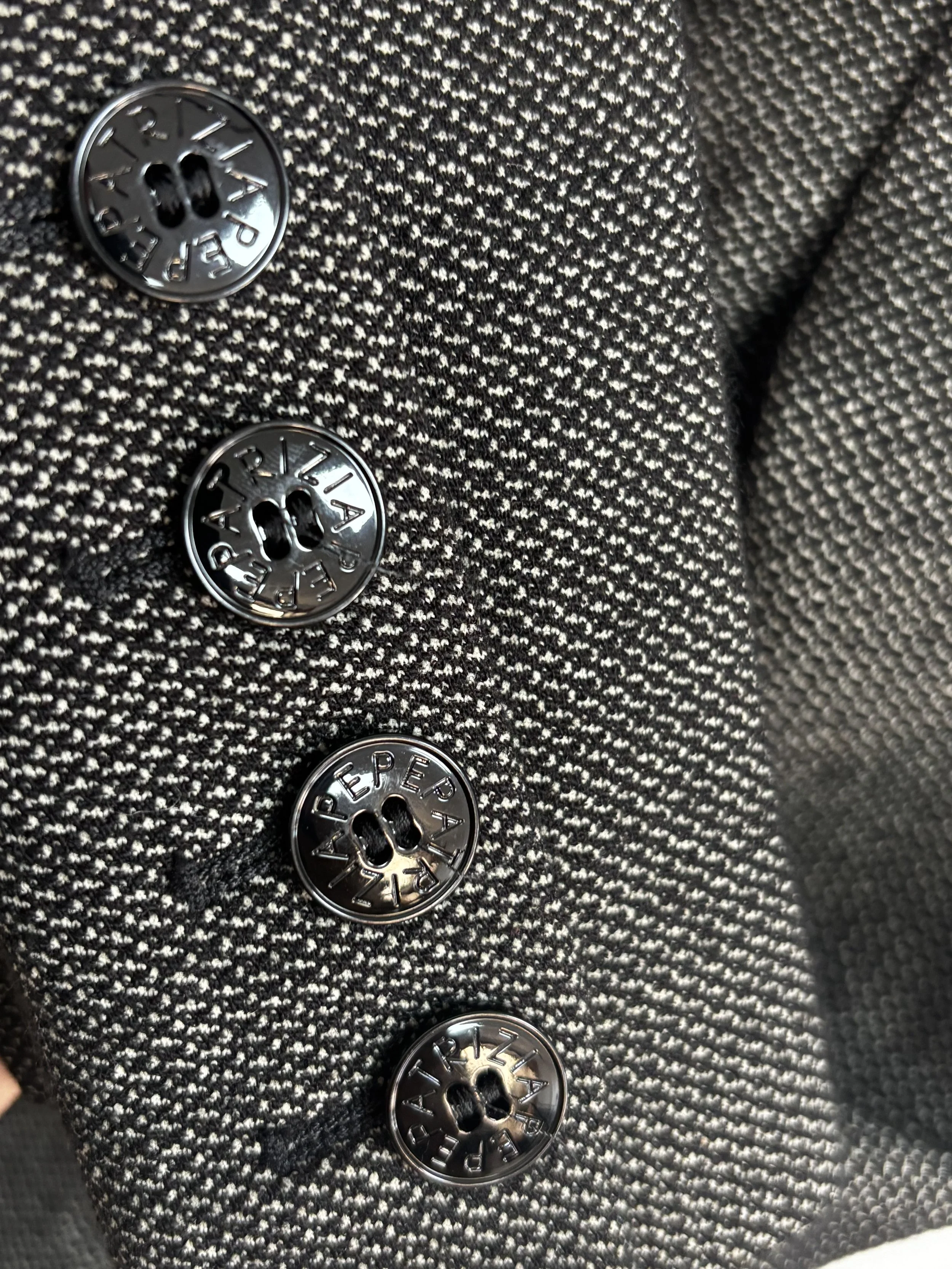 Macro detail of four Patrizia Pepe engraved buttons on a grey textured designer blazer sleeve, highlighting the branded hardware and fabric weave.