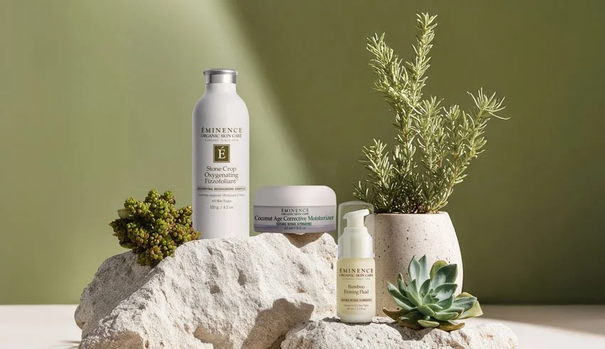 Skincare products including a bottle of Owen + Lauren Stone Crop Oxygenating Exfoliant, a jar of Eminence Coconut Age Corrective Moisturizer, a bottle of Eminence Bamboo Firming Fluid, and a potted plant with green foliage, arranged on rocks against a green and white background.