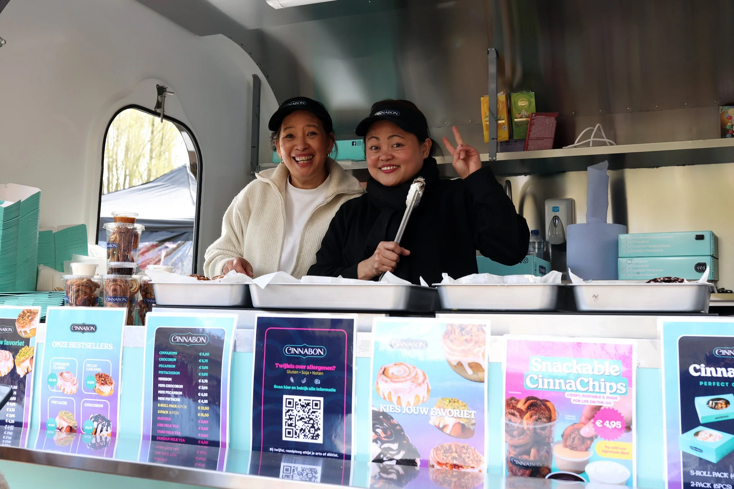 Twee glimlachende Cinnabon-medewerkers serveren klanten vanuit de Cinnabon on Wheels foodtruck in het Bloesempark tijdens het paasweekend