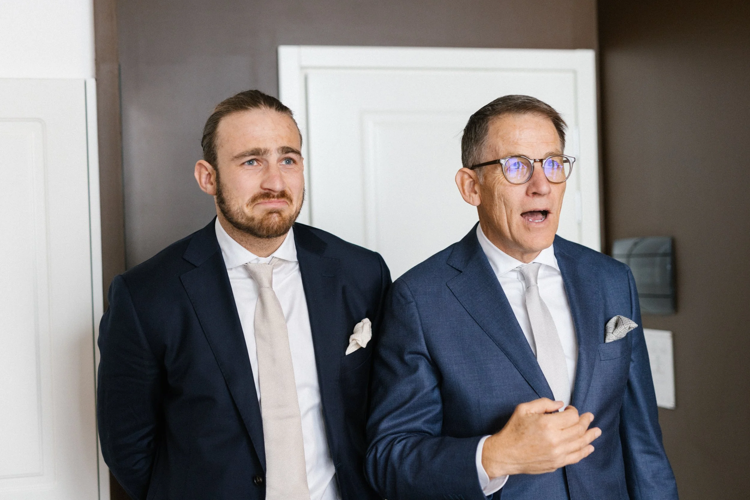 Two men in suits, one younger with a distressed expression and the other older with glasses, speaking or addressing a crowd.