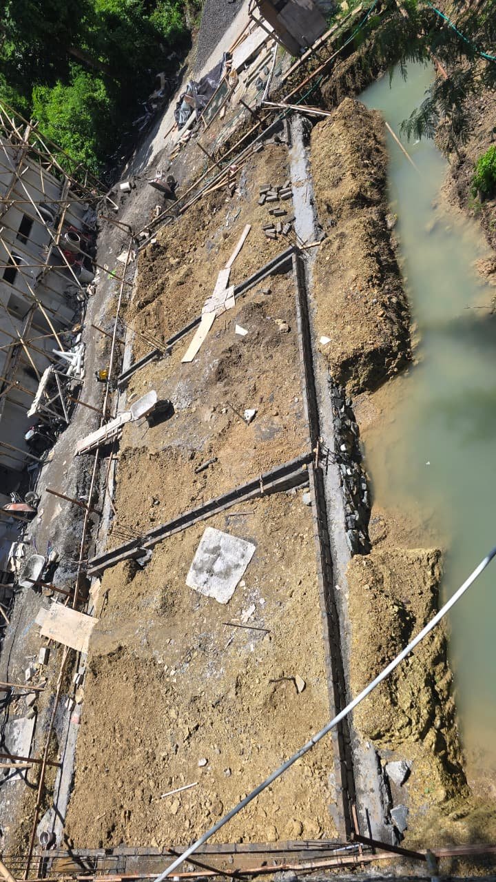 Construction site with exposed soil, concrete foundation, and pipes along a riverbank during daylight.