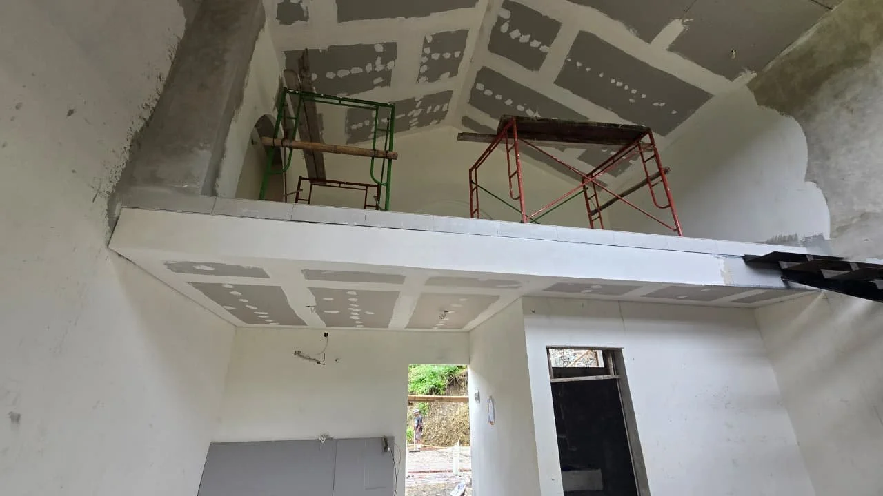 Interior of a building under construction, with scaffolding on a mezzanine level and unpainted drywall on the ceiling and walls.