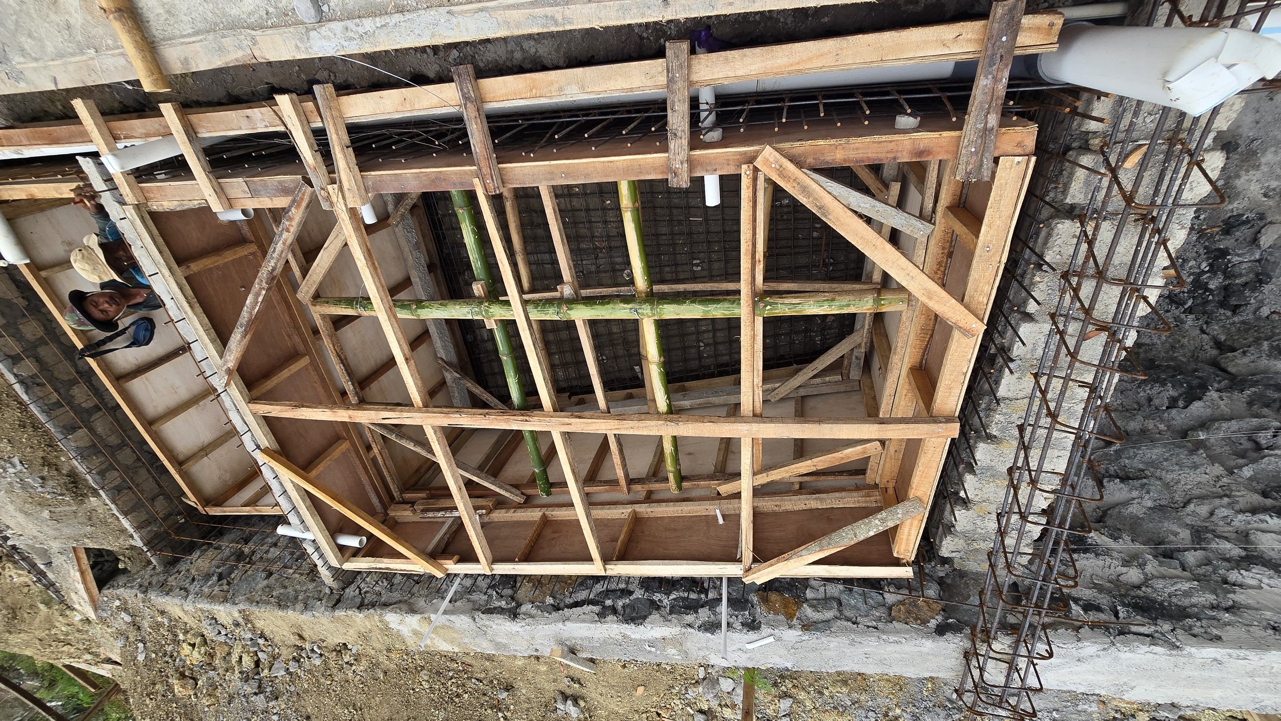 Construction site with wooden framing, bamboo supports, steel rebar, and workers in the background.