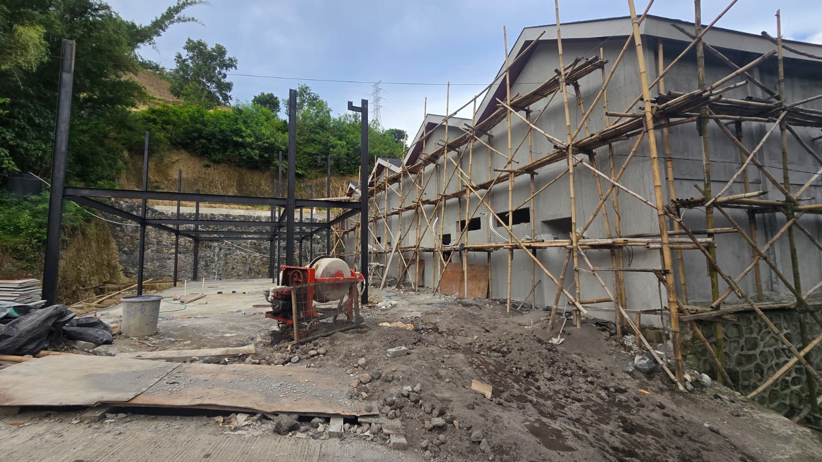 Construction site with a partially built concrete building covered in bamboo scaffolding, construction tools, and debris on the ground, surrounded by green trees and a rocky hillside.