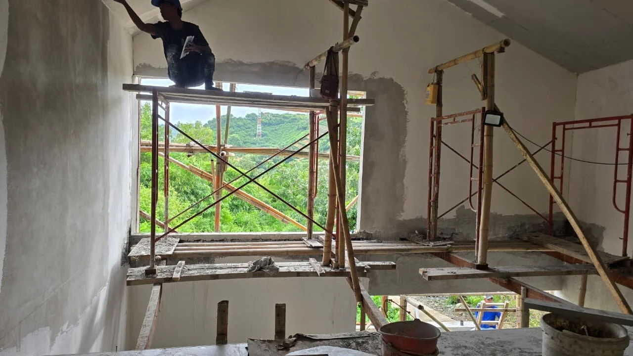 Construction worker on scaffolding inside a building, working near a large window opening, with green trees visible outside.
