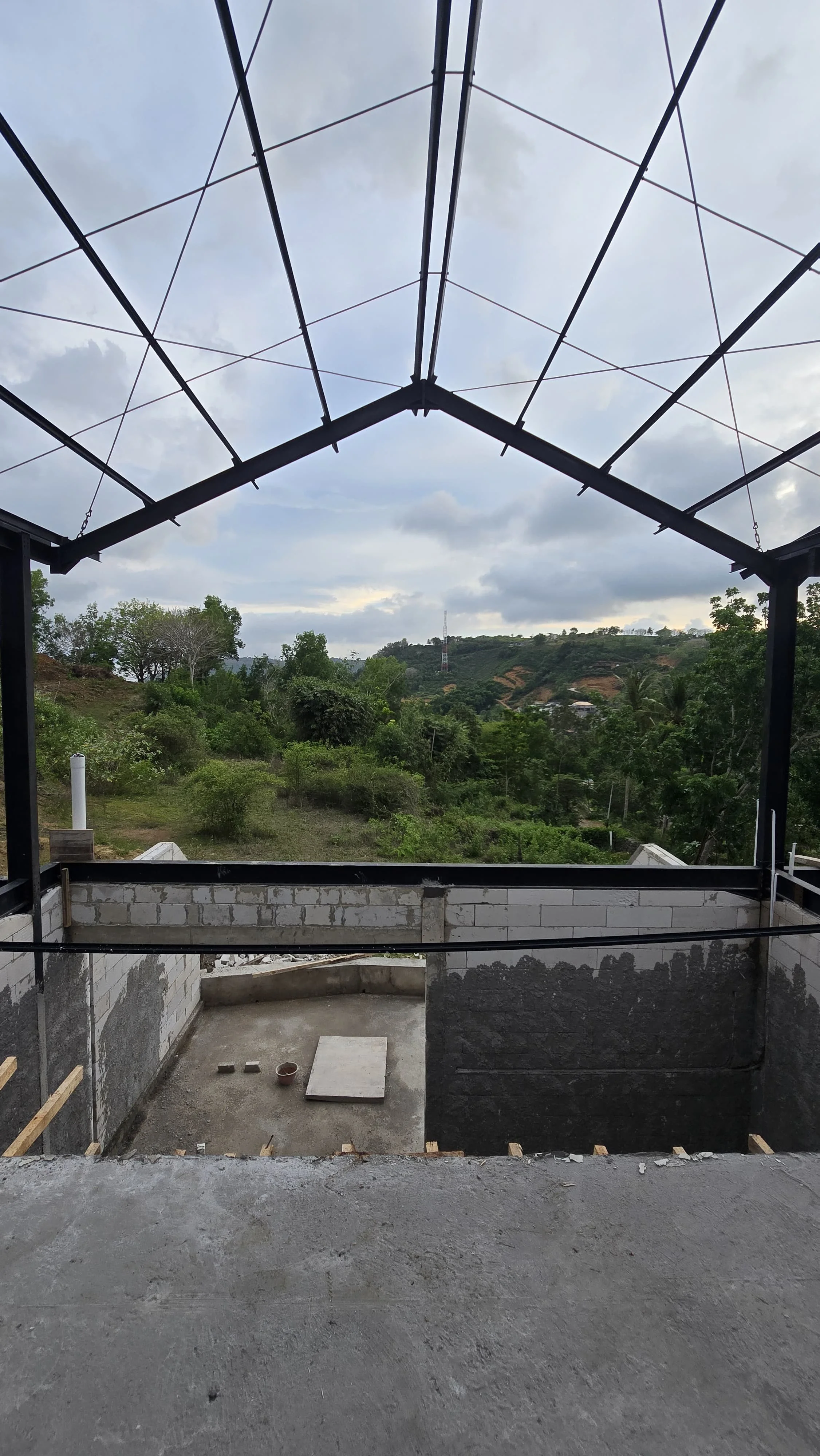 Construction site with a view of green hills and cloudy sky, partially built structure with concrete floor, brick walls, and metal framework.