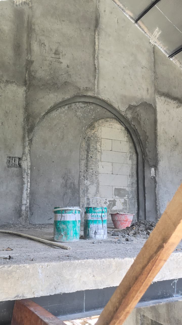 Interior view of an unfinished building with a brick arched wall opening. Construction supplies, including buckets and a red container, are on the floor. The wall is made of concrete blocks and appears to be incomplete.