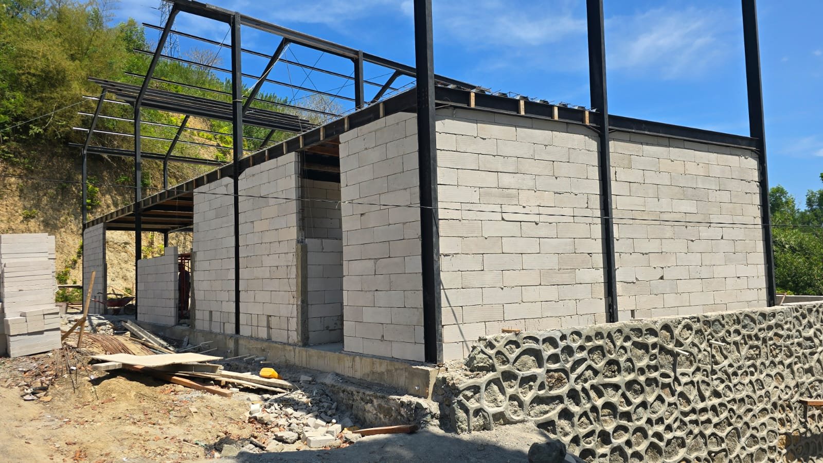 Construction site with partially built brick walls, black steel framework, and a decorative stone wall at the base. Clear blue sky and greenery in the background.