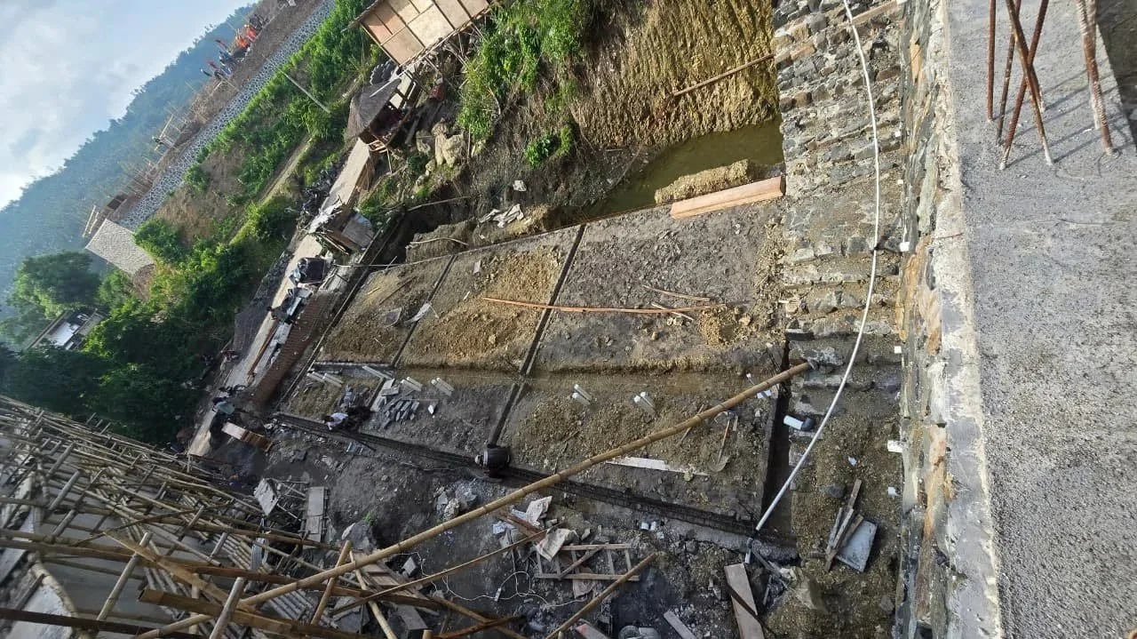 Construction site with exposed foundation, construction materials, and machinery, against a backdrop of trees and hills.