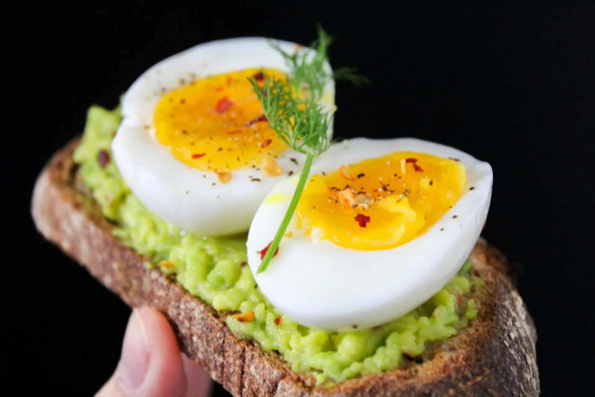 Close-up of an open-faced sandwich with mashed avocado, halved soft-boiled eggs, and garnished with herbs and red pepper flakes on dark background.