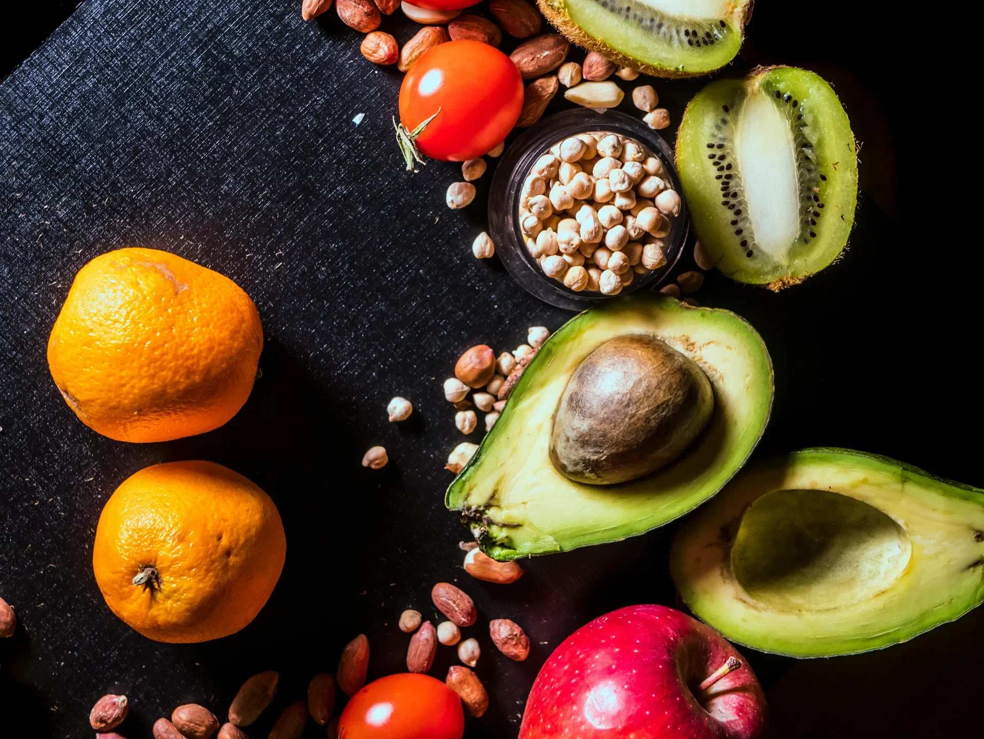 A variety of fresh fruits including avocados, kiwis, tomatoes, oranges, apples, and scattered nuts on a black surface.