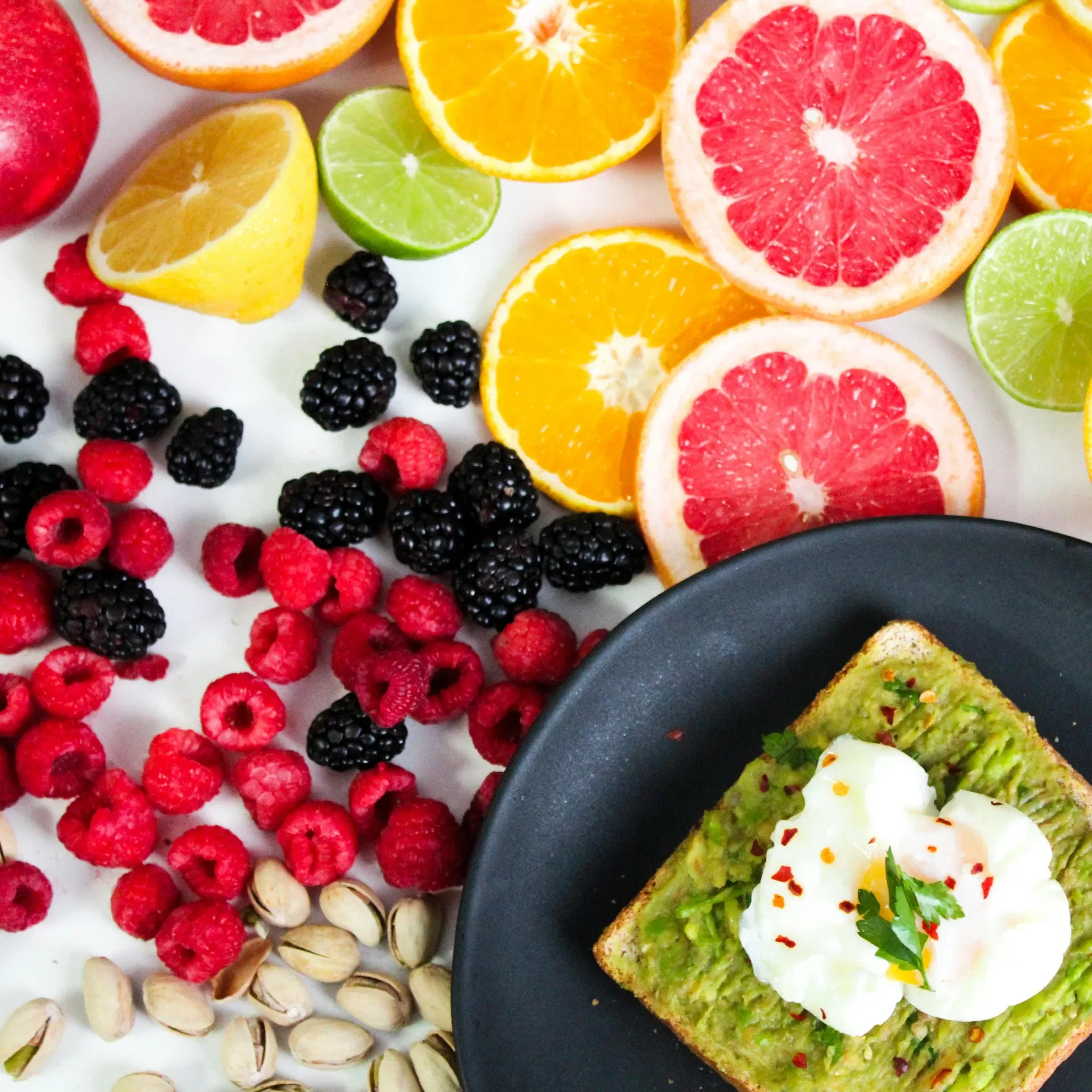A variety of sliced citrus fruits including grapefruits, oranges, lemons, and limes, arranged on a white surface, with a black plate holding avocado toast topped with a poached egg, red pepper flakes, and cilantro.