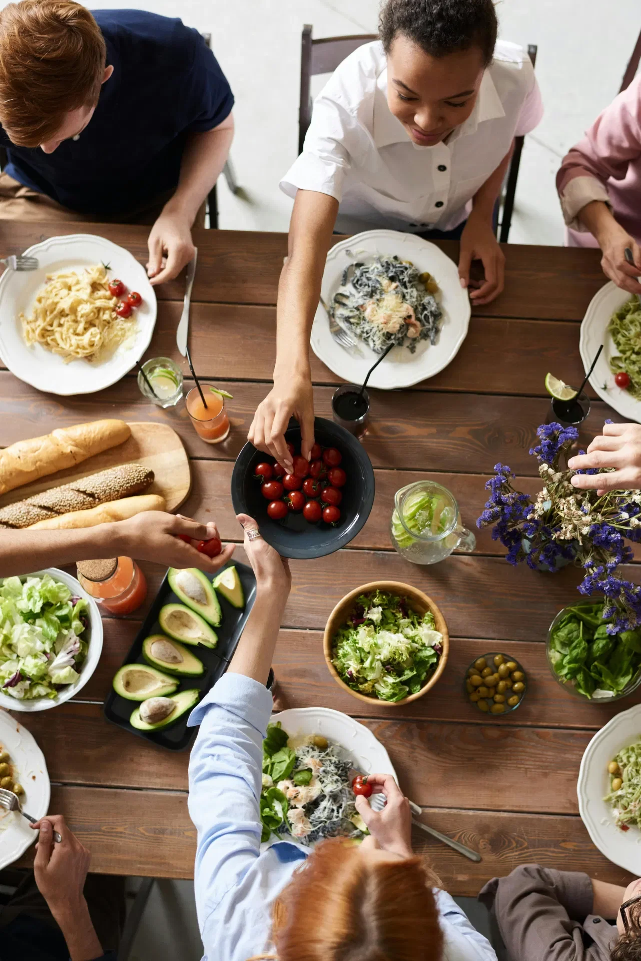 People gathering around a wooden table with plates of pasta, salads, bread, cherry tomatoes, avocados, olives, and drinks, sharing a meal.