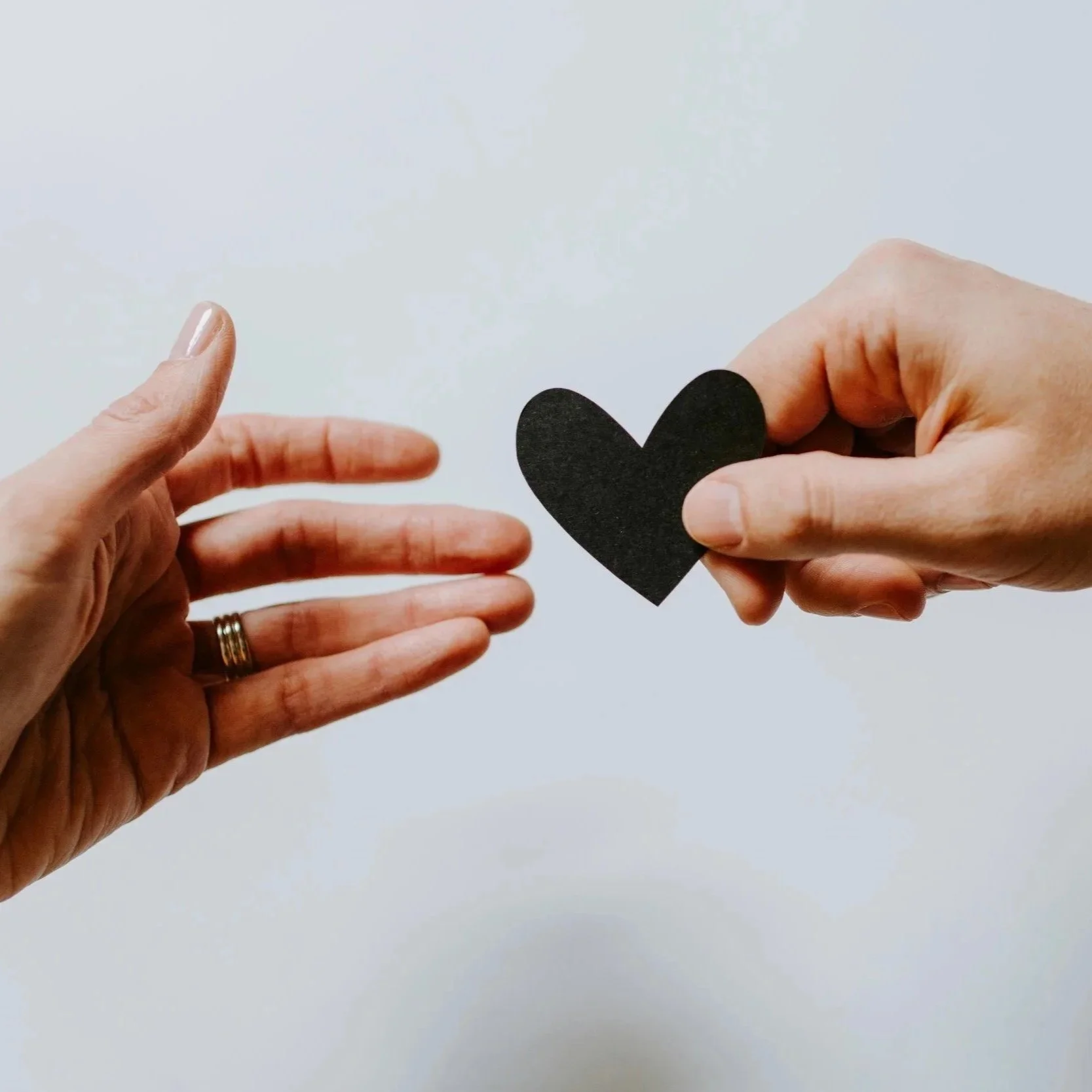 Two hands, one holding a black paper heart and the other reaching out, against a white background.