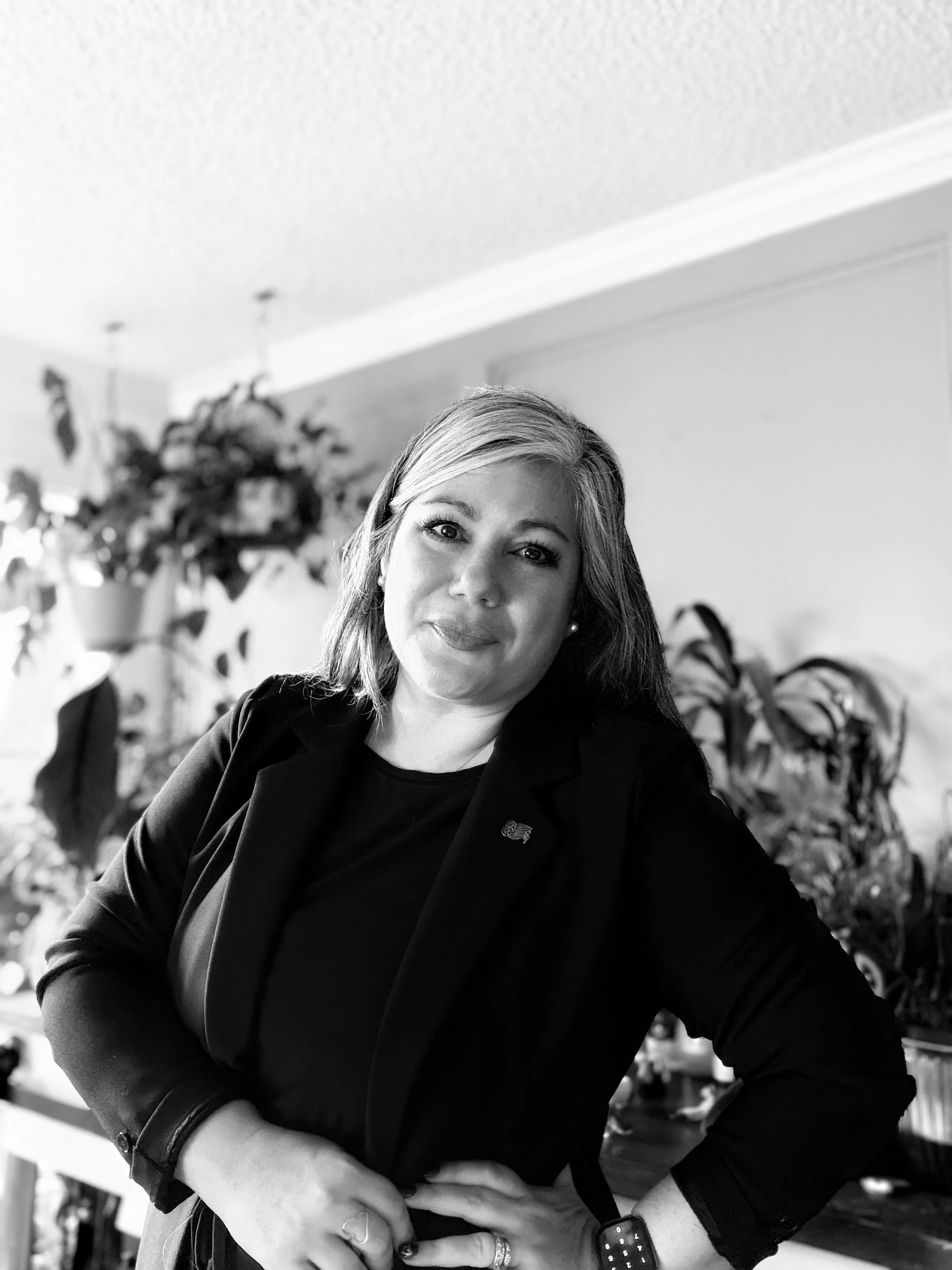 A woman with shoulder-length hair, wearing a blazer and a watch, smiling and posing indoors, with houseplants and a shelf in the background.
