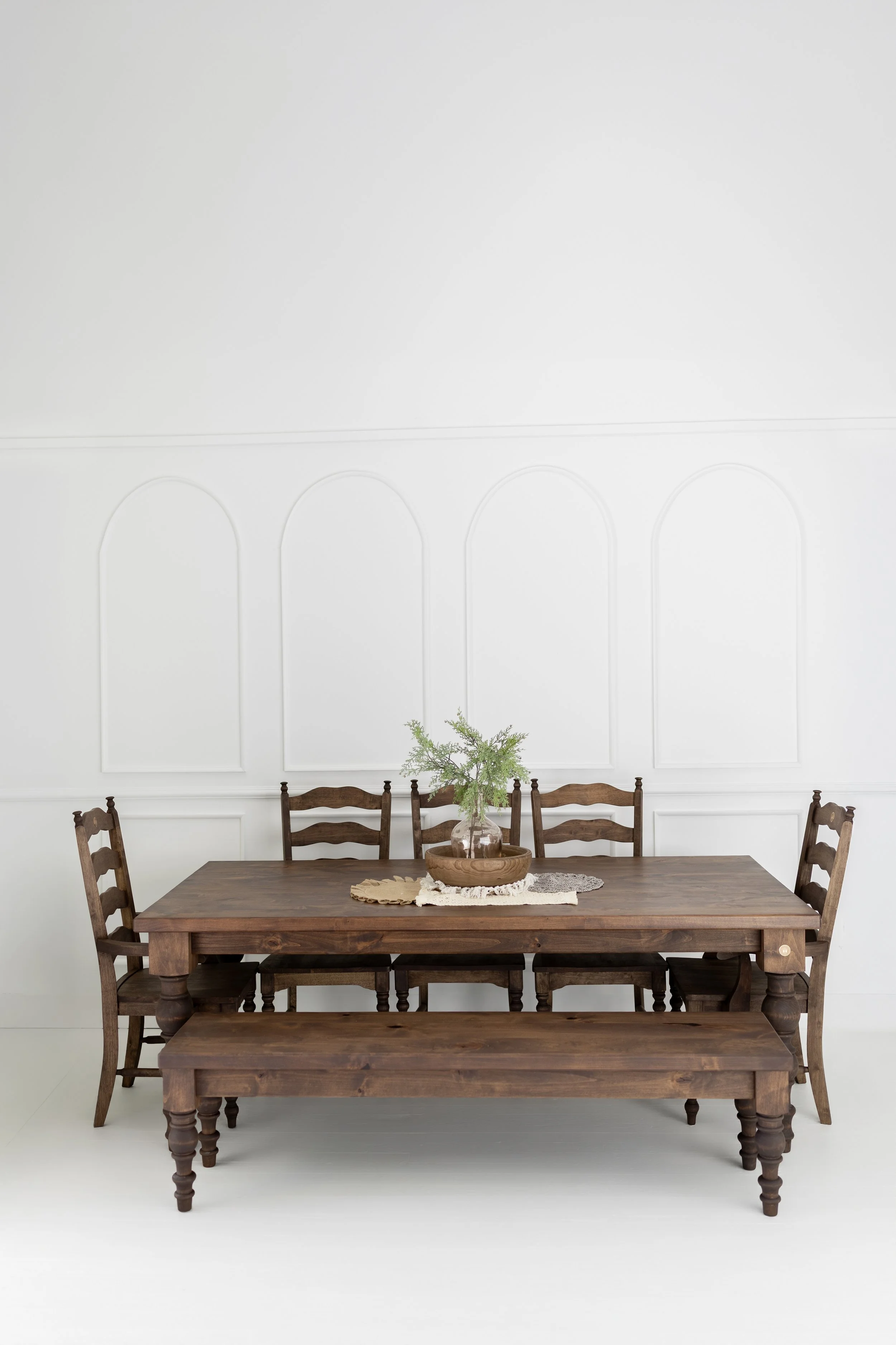 A wooden dining table with chairs, a vase with green plant, and a lace doily on a white background with decorative molding on the wall.