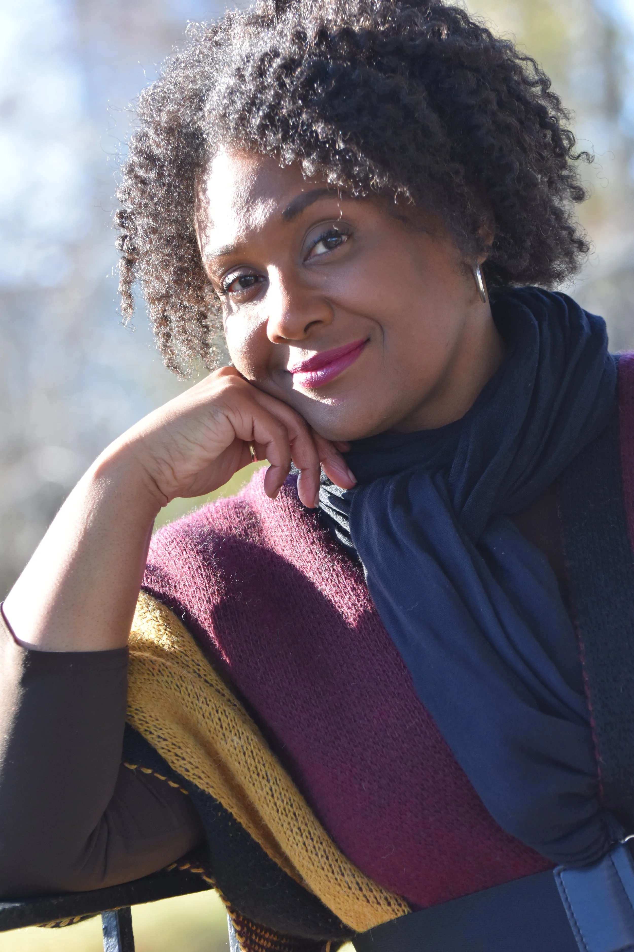 A woman with curly hair wearing a maroon and yellow sweater, black long sleeve shirt, and a navy blue scarf, smiling and resting her chin on her hand outdoors.