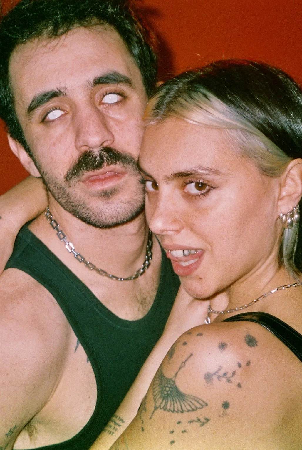 Un homme et une femme prennent un selfie dans une ambiance colorée, tous deux avec un style punk ou gothique, portant des bijoux en chaîne, tatouages visibles et une expression provocante.