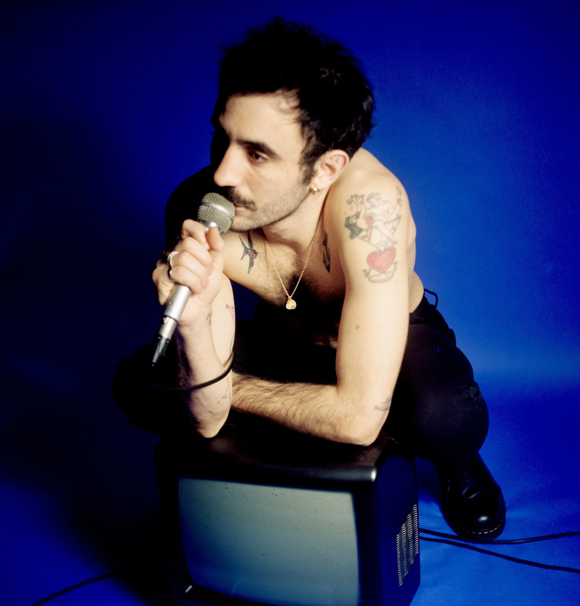 Un homme tatoué en train de chanter dans un micro, assis sur un meuble, contre un fond bleu.