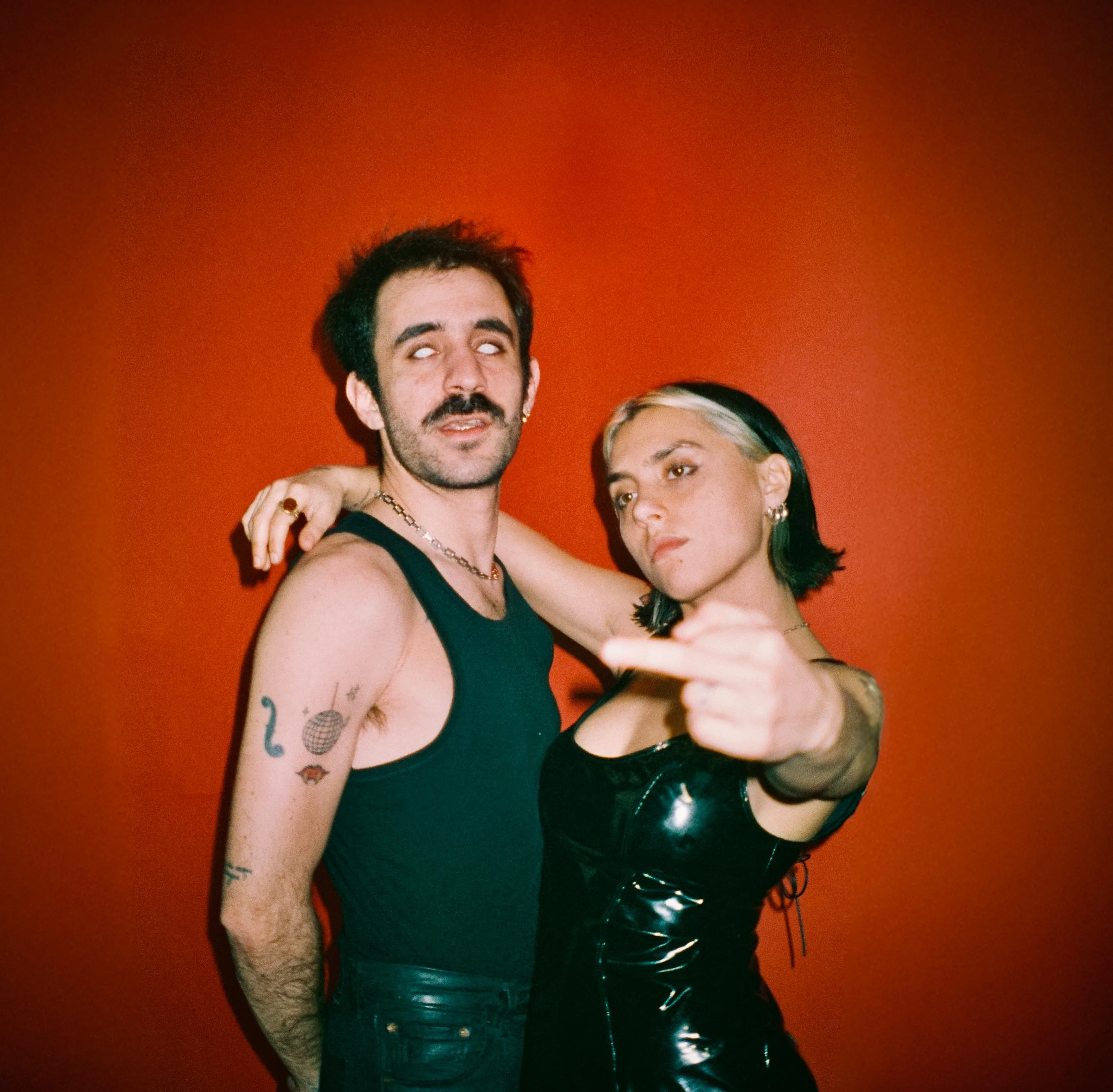 Un homme et une femme posent contre un mur rouge. L'homme porte un débardeur noir et a des tatouages, l'homme a une barbe, une moustache et des cheveux noirs. La femme porte un vêtement noir en cuir, avec des cheveux noirs avec une mèche blonde. Elle