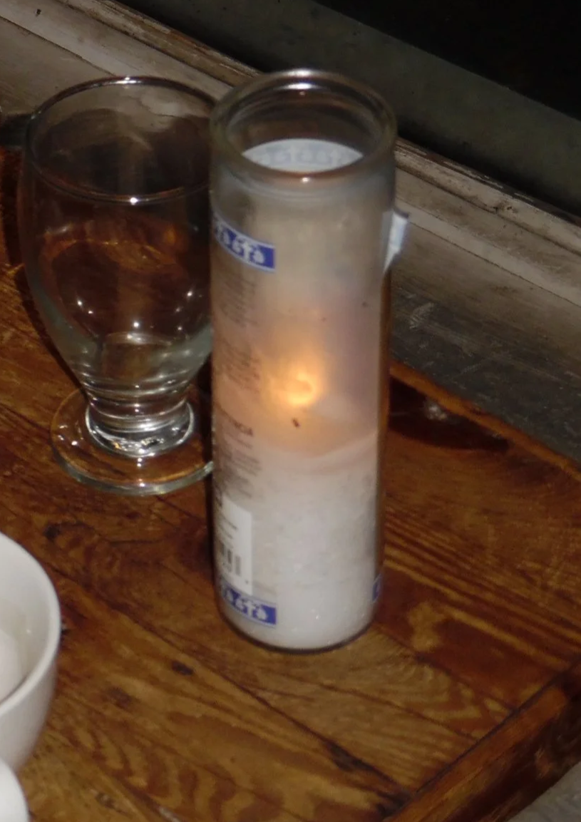 A lit white candle in a clear glass container next to an empty glass on a wooden table.