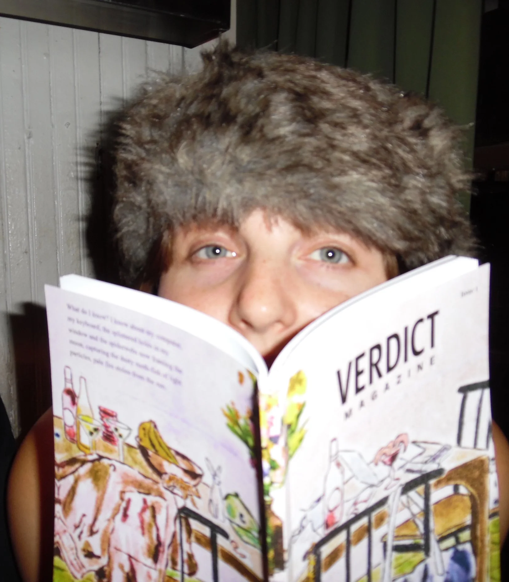 A person wearing a furry winter hat reading a magazine called "Verdict Magazine".